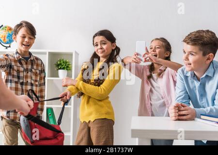 Selektiver Fokus des grausamen Schulkindes mit Smartphone in der Nähe von Klassenkameraden beim Mobbing von Schulmädchen Stockfoto