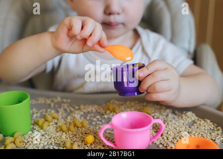 Kleines Kind gießt den gelben Löffel des Spielzeugs voll in der Tasse. Spiele mit Produkten. Montessori-Technik: Entwicklung von guten motorischen Fähigkeiten bei Kindern Stockfoto