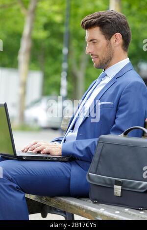 Der Geschäftsmann, der seinen Laptop benutzt, während er auf der Bank sitzt Stockfoto