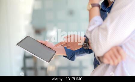 Seitenansicht von zwei Geschäftsleuten, die sich mit einem Mock-up-Tablet beraten, während sie an ihrem Arbeitsplatz stehen Stockfoto