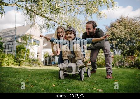 Junge, die auf dem Spielzeugauto sitzen und so vorgeben zu fliegen, Eltern ermutigen ihn Stockfoto