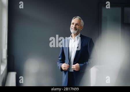 Porträt eines lächelnden, reifen Geschäftsmannes, der im Büro einen blauen Anzug trägt und auf Distanz blickt Stockfoto