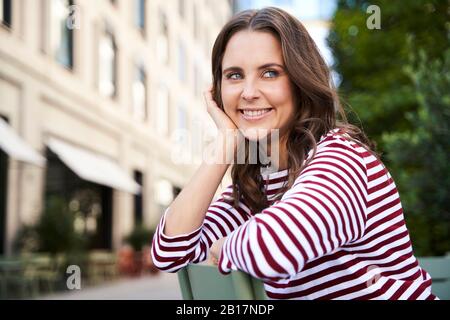 Porträt von lächelnden Brünette Frau auf einem Stuhl in der Stadt sitzen Stockfoto