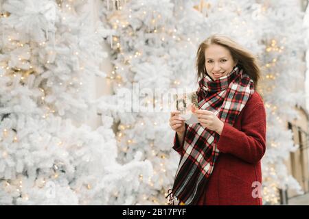 Porträt einer lächelnden jungen Frau, die auf der Straße steht, mit Weihnachtskarte vor weißen Weihnachtsbäumen Stockfoto