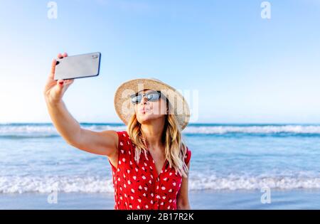 Blonde Frau trägt rotes Kleid und Hut und mit Smartphone und ein Selfie am Strand, einen küssenden Mund zu machen Stockfoto