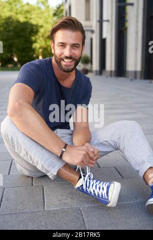 Porträt eines lächelnden Mannes, der auf dem Boden in der Stadt sitzt und seine Schuhe schnürt Stockfoto