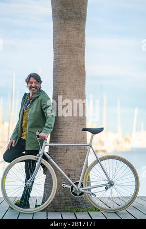 Portrait des lachenden reifen Mannes mit fixie Fahrrad gegen Palmenstamm lehnt, Alicante, Spanien Stockfoto
