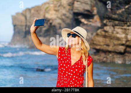 Blonde Frau in rotem Kleid und Hut und mit Smartphone und ein Selfie am Strand Stockfoto