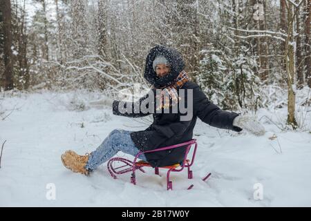 Lächelnde Frau auf Schlitten sitzen Schnee in die Luft im Winterwald werfen Stockfoto