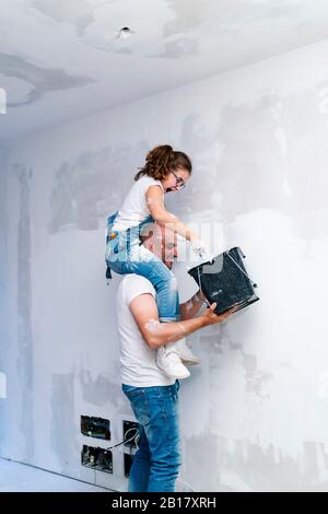 Mädchen, die auf den Schultern des Vaters sitzen und die Wand ihres neuen Hauses malen Stockfoto