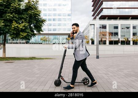 Legerer junger Geschäftsmann mit elektrischem Roller telefoniert in der Stadt Stockfoto