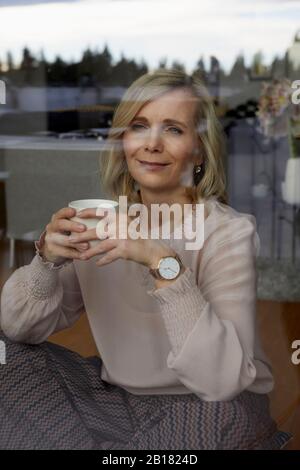 Lächelnde blonde Frau hält Kaffeetasse hinter Fensterscheibe zu Hause Stockfoto