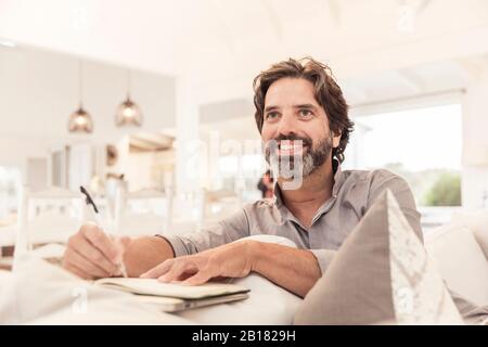 Lächelnder bärtiger Mann, der auf der Couch sitzt und Notizen nimmt Stockfoto