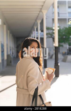 Porträt einer lächelnden jungen Frau mit Buch und Kaffee zum Mitnehmen in der Stadt Stockfoto