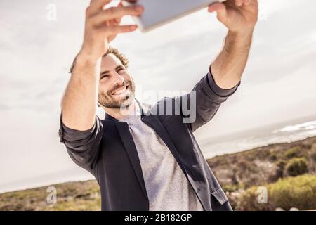 Fröhlicher junger Mann in Business-Jacke, der ein Selfie an der Küste macht Stockfoto