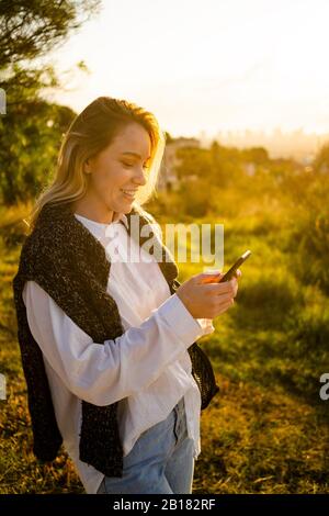 Lächelnde junge Frau mit Handy bei Sonnenaufgang in der Natur Stockfoto