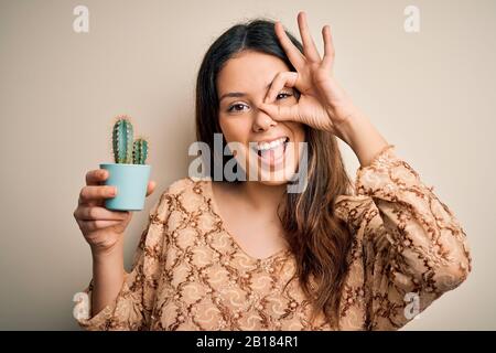 Junge schöne Brunette-Frau, die Kaktuskanne über isoliertem weißem Hintergrund hält, mit fröhlichem Gesicht lächelnd, mit einem guten Schild, das mit der Hand auf dem Auge durch die Augen blickt Stockfoto