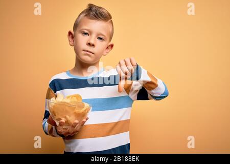 Junge kleine kaukasische Kinder, die unerhitzte Kartoffeln essen, knacken Pommes über gelben Hintergrund mit wütenden Gesicht, negatives Zeichen, das mit Daumen ablehnt Stockfoto