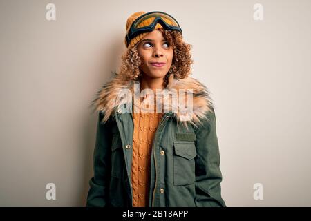 Junge afroamerikanische Skifahrerin mit lockigen Haaren, die Schneesportkleidung und eine Skibrille trägt, lächelt lächelnd auf die Seite und starrt wegdenkend. Stockfoto