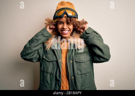 Junge afroamerikanische Skifahrerin mit lockigen Haaren, die Schneesportkleidung trägt, und Skihoggles, Die Mit den Fingern an den Ohren ziehen, lustige Geste. Hörspiel Stockfoto
