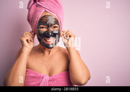 Frau im mittleren Alter, die schöne schwarze Gesichtsmaske über isoliertem rosafarbenem Hintergrund Trägt, Lächelnde Ohren mit Fingern ziehen, lustige Geste. Hörprobe Stockfoto