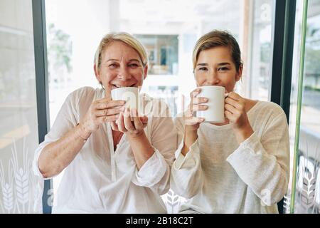 Glücklich lächelnde, reife Frau und ihre hübsche Erwachsene Tochter trinken große Tassen Morgenkaffee und lächeln mit der Kamera Stockfoto