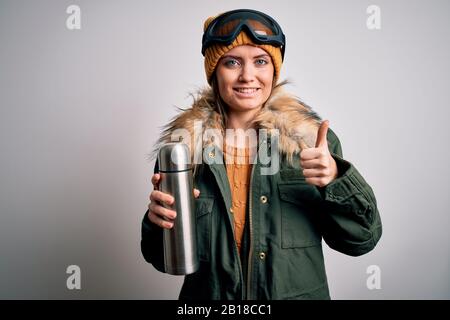 Junge, schöne Skifahrerin mit blauen Augen, die eine Skibrille trägt, die Thermo mit Kaffee trinkt, glücklich mit einem großen Lächeln, mit einem guten Schild, Daumen nach oben mit den Fingern, Stockfoto