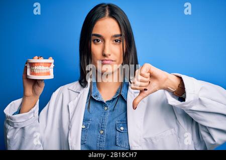 Junge schöne brünette Zahnärztin, die Mantel trägt, die Kunststoff-Zahnprothese mit verärgertem Gesicht hält, negatives Zeichen, das mit Daumen nach unten ablehnt, Stockfoto