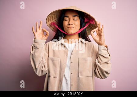Junge schöne Frau mit traditionellem, konischen asiatischen Hut über isoliertem rosafarbenem Hintergrund entspannen Sie sich und lächeln Sie mit geschlossenen Augen, die Meditationsgeste ausführen Stockfoto