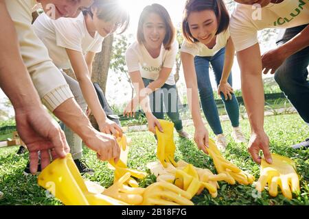 Lächelnde asiatische Freiwillige, die gelbe Gummihandschuhe ansetzen, bevor sie mit der Reinigung des Parks beginnen und Müll sammeln Stockfoto