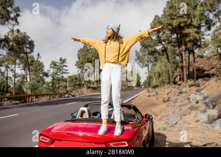 Porträt einer jungen, stilvollen Frau mit einem hellen Pullover, der mit dem Auto unterwegs ist, schöne Landschaften genießt und die Hände am Straßenrand im Wald erhöht. Sorgloses Lifestyle- und Reisekonzept Stockfoto