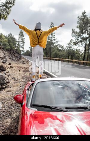 Junge, stilvolle Frau, die mit dem Auto unterwegs ist und dabei schöne Landschaften genießt, während sie am Straßenrand steht, Blick auf die Rückseite. Sorgloses Lifestyle- und Reisekonzept Stockfoto