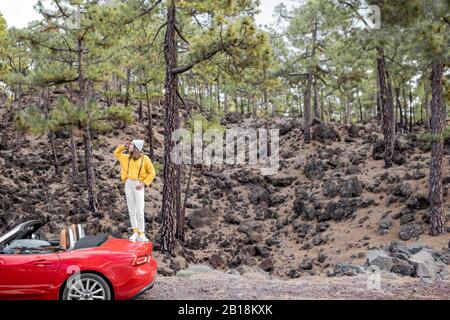 Junge, stilvolle Frau, die mit dem Auto unterwegs ist und schöne Landschaften genießt, während sie am Straßenrand im Wald steht. Sorgloses Lifestyle- und Reisekonzept Stockfoto