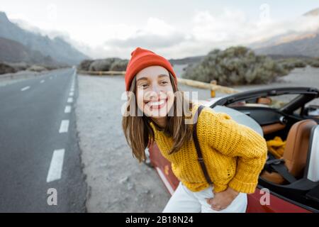 Porträt einer fröhlichen Frau, die mit einem hellen Pullover und einem Hut bekleidet ist, die eine Fahrt genießen, in der Nähe des Autos am Straßenrand des malerischen Vulkantals bei Sonnenuntergang Stockfoto