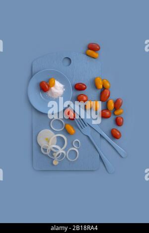 Rote und gelbe Kirschtomaten auf blauem Teller, blaues Messer, blaue Gabel, auf blauem Grund.Stillleben. Vertikal. Die Ansicht von oben. Geeignet als Stockfoto