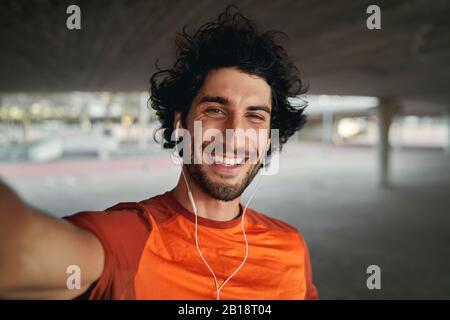 Porträt eines lächelnden jungen Mannes mit Ohrhörern in den Ohren, der selfie draußen nimmt - Foto eines Mannes, der lächelnd die Kamera anschaut Stockfoto