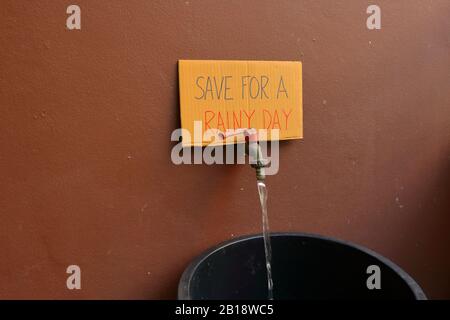 Closeup Phrase SAVE FOR A REGNERY DAY written on pappe sign placed on faucet with water flowing into big Black Bucket Stockfoto