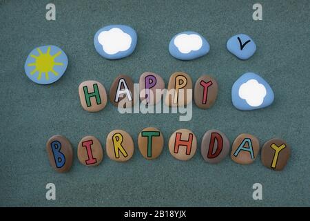 Kreative Botschaft für einen Glücklichen Geburtstag mit einer Komposition aus farbigen Steinbuchstaben mit Sonne und Wolken gestalten sich über grünem Sand Stockfoto
