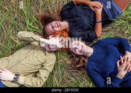 Sommerferien Urlaub glückliche Menschen Konzept. Draufsicht Gruppe von drei Freunden, die im Kreis auf Gras liegen, lächeln und zusammen im Freien Spaß haben. Picknick mit Freunden auf dem Weg in die Natur Stockfoto
