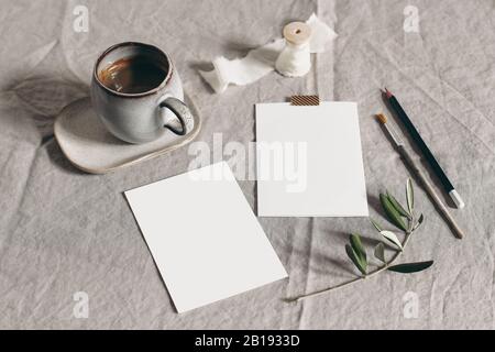 Moody feminine Hochzeitstatikerei Mock-up-Szene. Leere Grußkarten, eine Tasse Kaffee und grüne olivgrüne Blätter, Äste auf Leinentischgrund Stockfoto