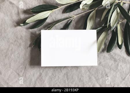 Nahaufnahme von leerem Gruß, Visitenkarte und grünen olivfarbenen Blättern, Ästen auf Leinentisch im Hintergrund. Moody feminine Hochzeitstatikerei Mock-up Stockfoto