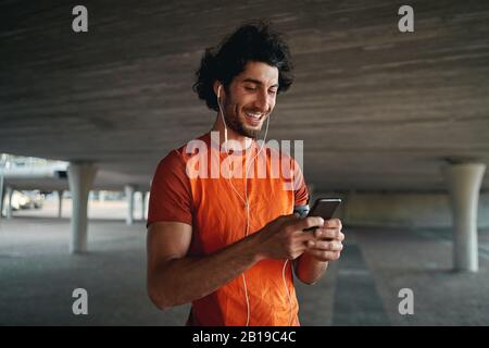 Porträt eines lächelnden jungen männlichen Läufers, der unter der Betonbrücke steht, mit Ohrhörer in den Ohren, der auf einem Smartphone tippt Stockfoto