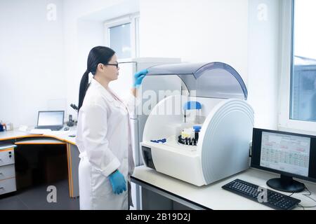 Dunkelhaariger Arzt in einer weißen Robe, die den hämatologischen Analysator öffnet Stockfoto