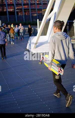 Ein Teenager mit einem Skateboard, der während des Sonnenuntergangs auf der Stadtbrücke spaziert Stockfoto