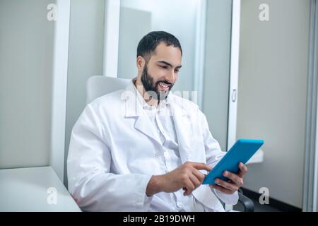 Männlicher Arzt in einer weißen Robe, der etwas liest und lächelt Stockfoto