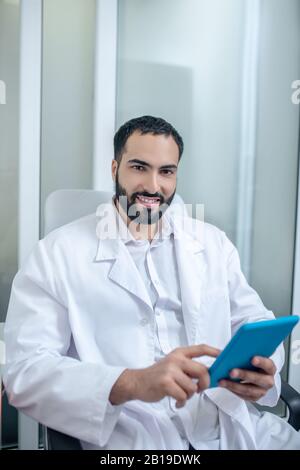 Männlicher Doktor in einer weißen Robe, die im Stuhl sitzt und Internet sucht Stockfoto
