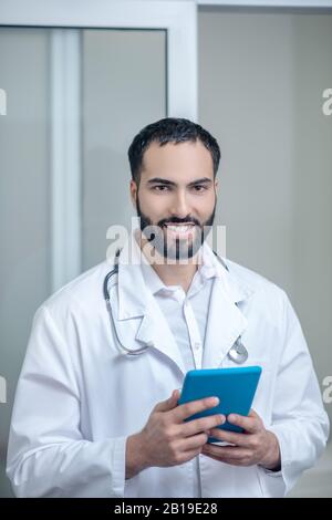 Männlicher Arzt in einer weißen Robe, die eine Tablette hält und lächelt Stockfoto