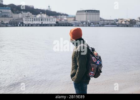 Junger bärtiger Reisender mit Flecken auf Rucksack an der Flussfront mit Stadtbild im Hintergrund. Stockfoto