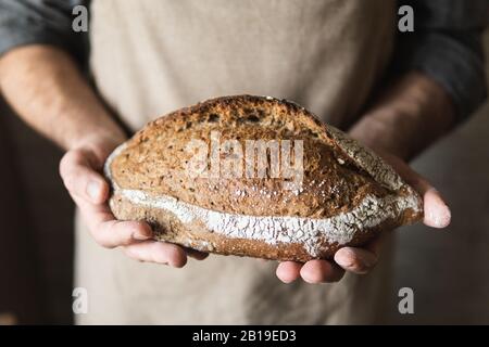Mann in rustikaler Schürze, der einen Brotlaib in den Händen hält. Nahaufnahme des Produkts. Stockfoto