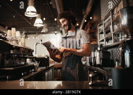 Fröhlicher junger kaukasischer Café-Besitzer, der schwarze gestreifte Schürze mit digitalem Tablet trägt Stockfoto